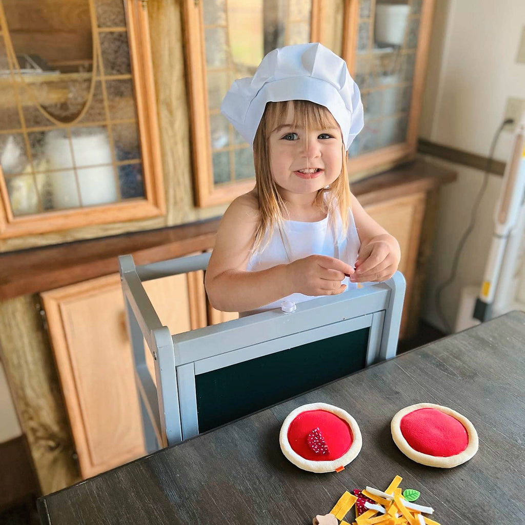 toddler girl wearing white chef hat and child's apron set while playing with pretend pizzas