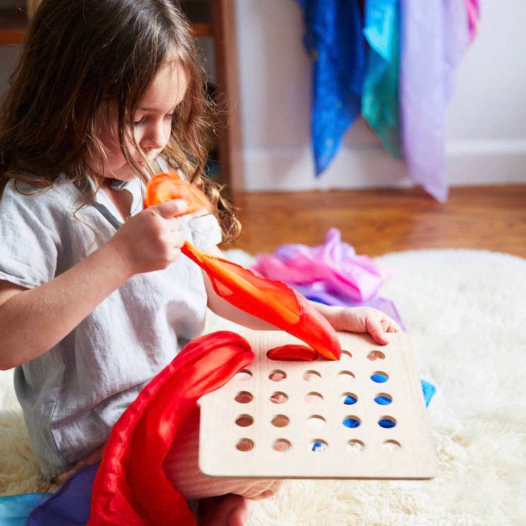 little girl weaving a silk scarf through a wooden Montessori sensory board